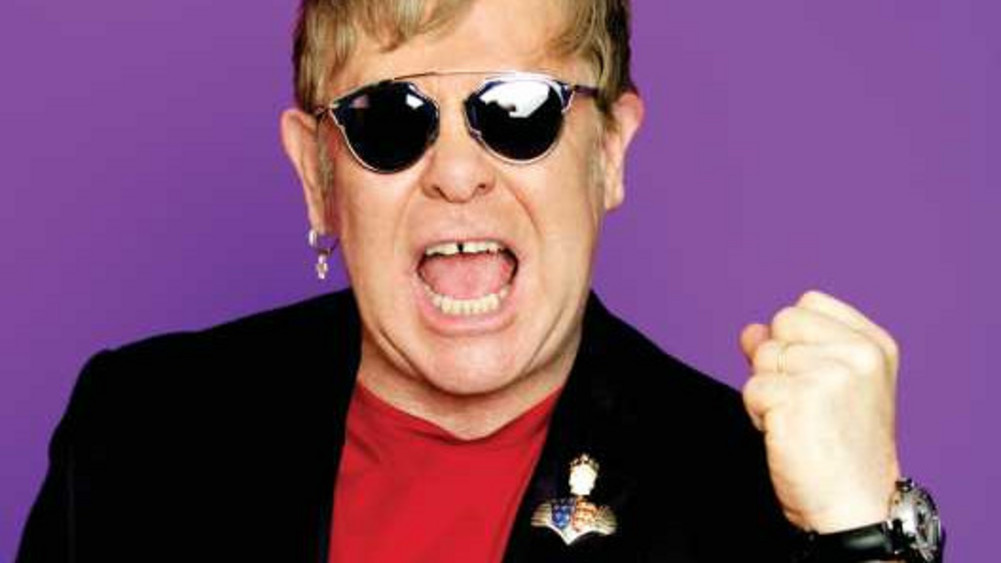 Elton John hat eine Sonnenbrille auf und hebt eine Faust, während er in die Kamera schreit