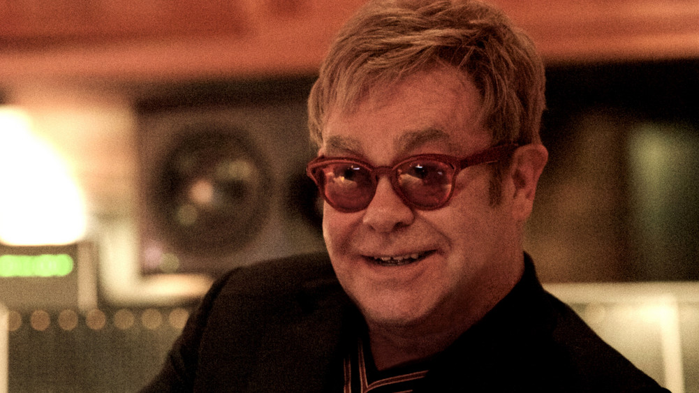 Elton John sitzt mit einer roten Brille in einem Studio