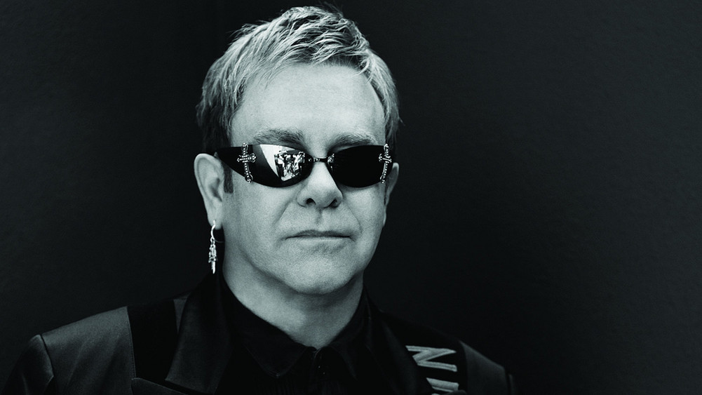 Elton John steht mit Sonnenbrille und Anzug vor dunklem Hintergrund